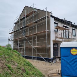 DAW- BUD - Budynek w trakcie prac elewacyjnych, widoczne rusztowania, częściowo ocieplona ściana i przenośna toaleta na pierwszym planie.