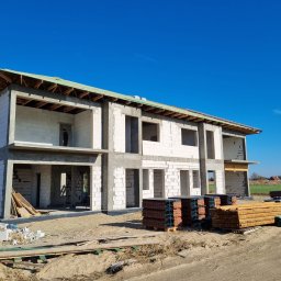 DAW- BUD - Surowy stan dwukondygnacyjnego domu z białych bloczków, widoczna konstrukcja dachu z zieloną membraną, stosy dachówek i drewna na placu budowy, błękitne niebo w tle.