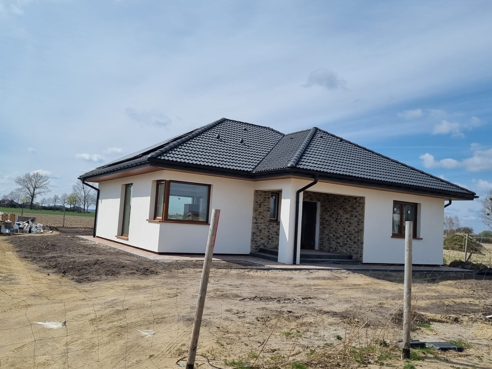 Nowoczesny, parterowy dom z białymi ścianami i ciemnoszarą dachówką, częściowo wykończony kamieniem elewacyjnym, na działce w trakcie zagospodarowywania.