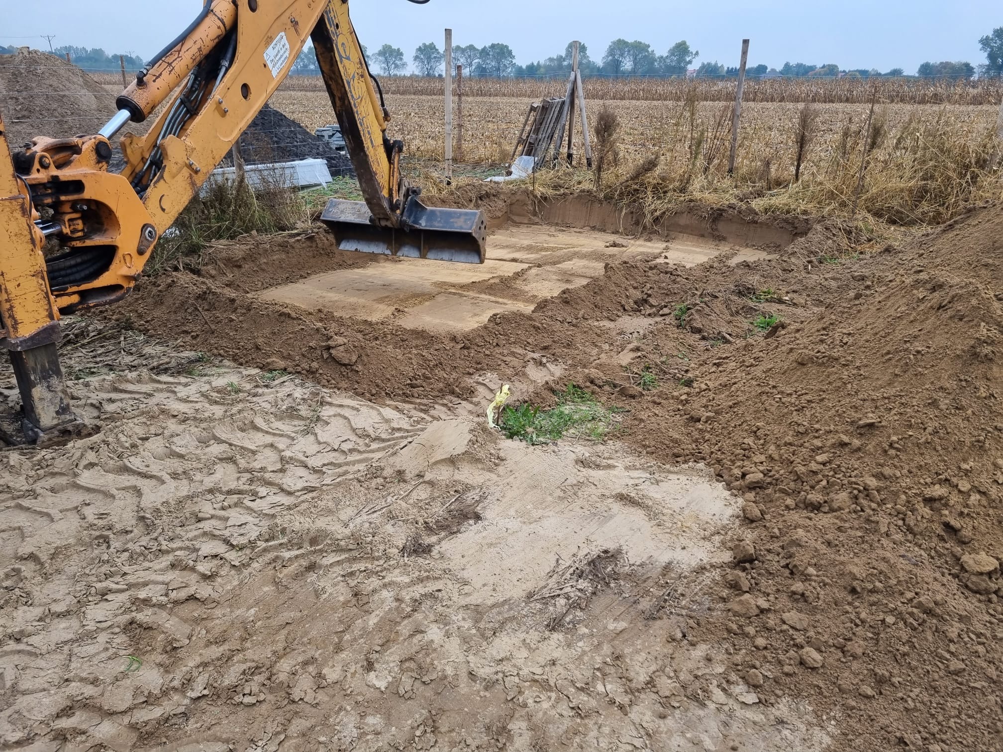 Żółta koparka przygotowuje wykop pod fundamenty na polu, widoczne ślady opon i wykopana ziemia, w tle ogrodzenie i pole kukurydzy.