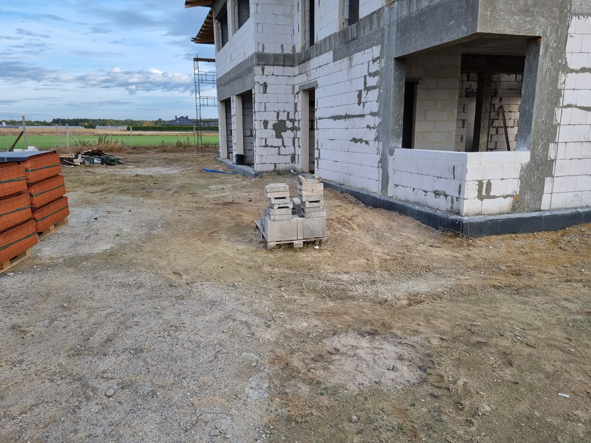 Surowy stan dwukondygnacyjnego domu w budowie z białych bloczków, widoczne rusztowanie i paleta z dachówką, teren budowy w trakcie prac.