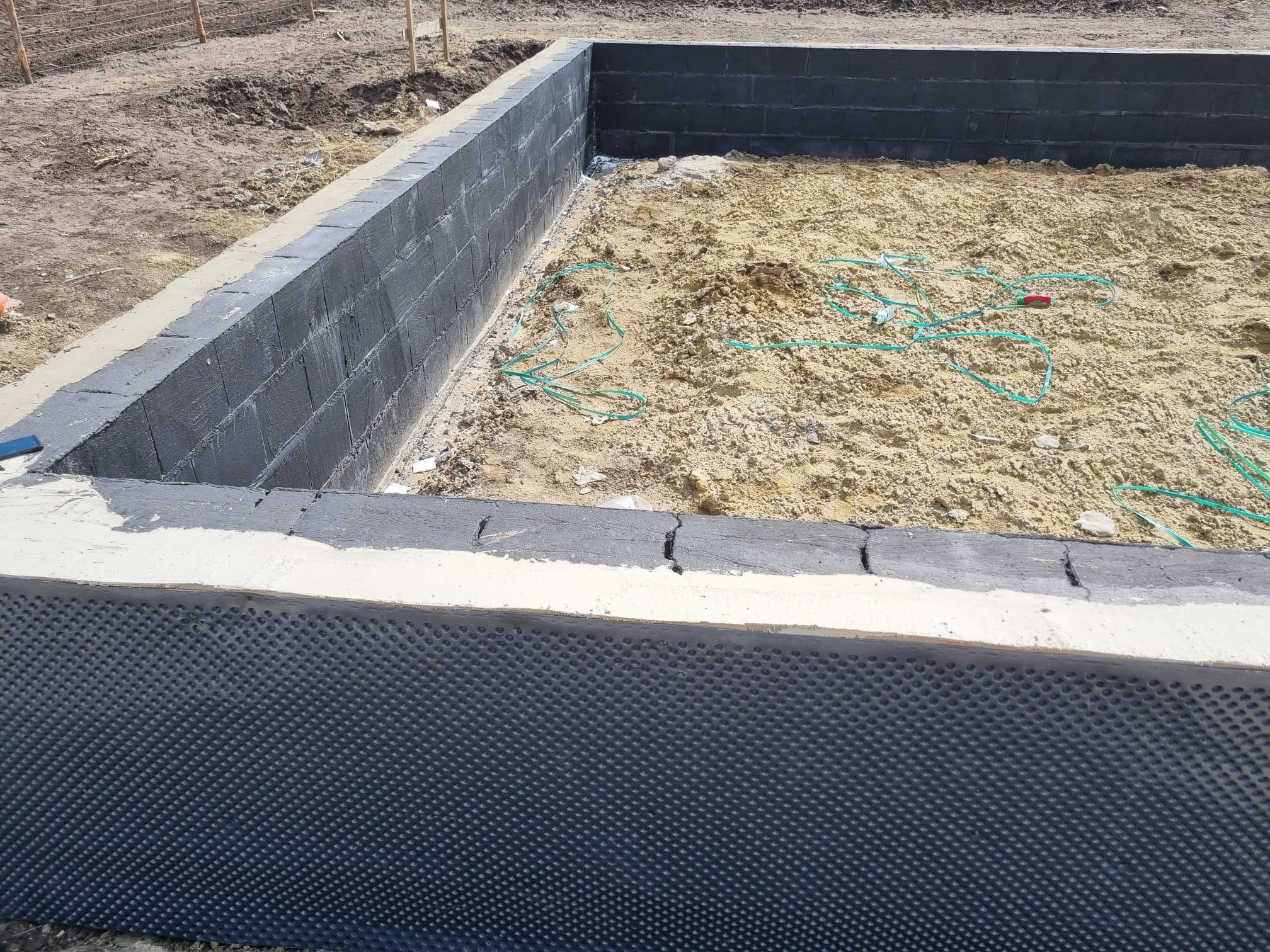 Wykop pod fundamenty z izolacją pionową w postaci folii kubełkowej, widoczne bloczki betonowe i piasek wewnątrz wykopu.