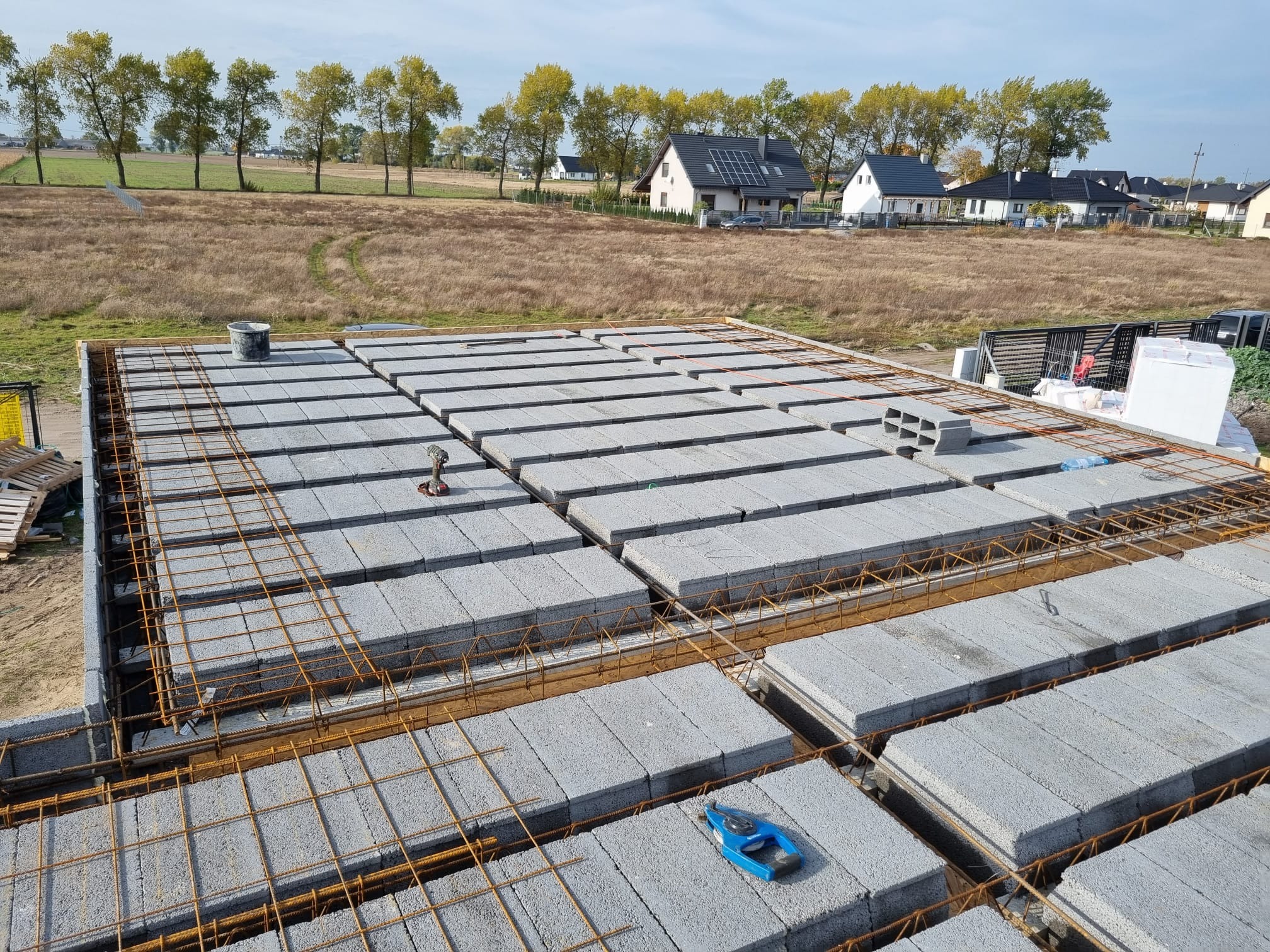 Wieniec stropowy z szalunkiem i zbrojeniem na budowie domu jednorodzinnego, widok z góry na pustaki szalunkowe i stalowe pręty, w tle zabudowa jednorodzinna i drzewa.