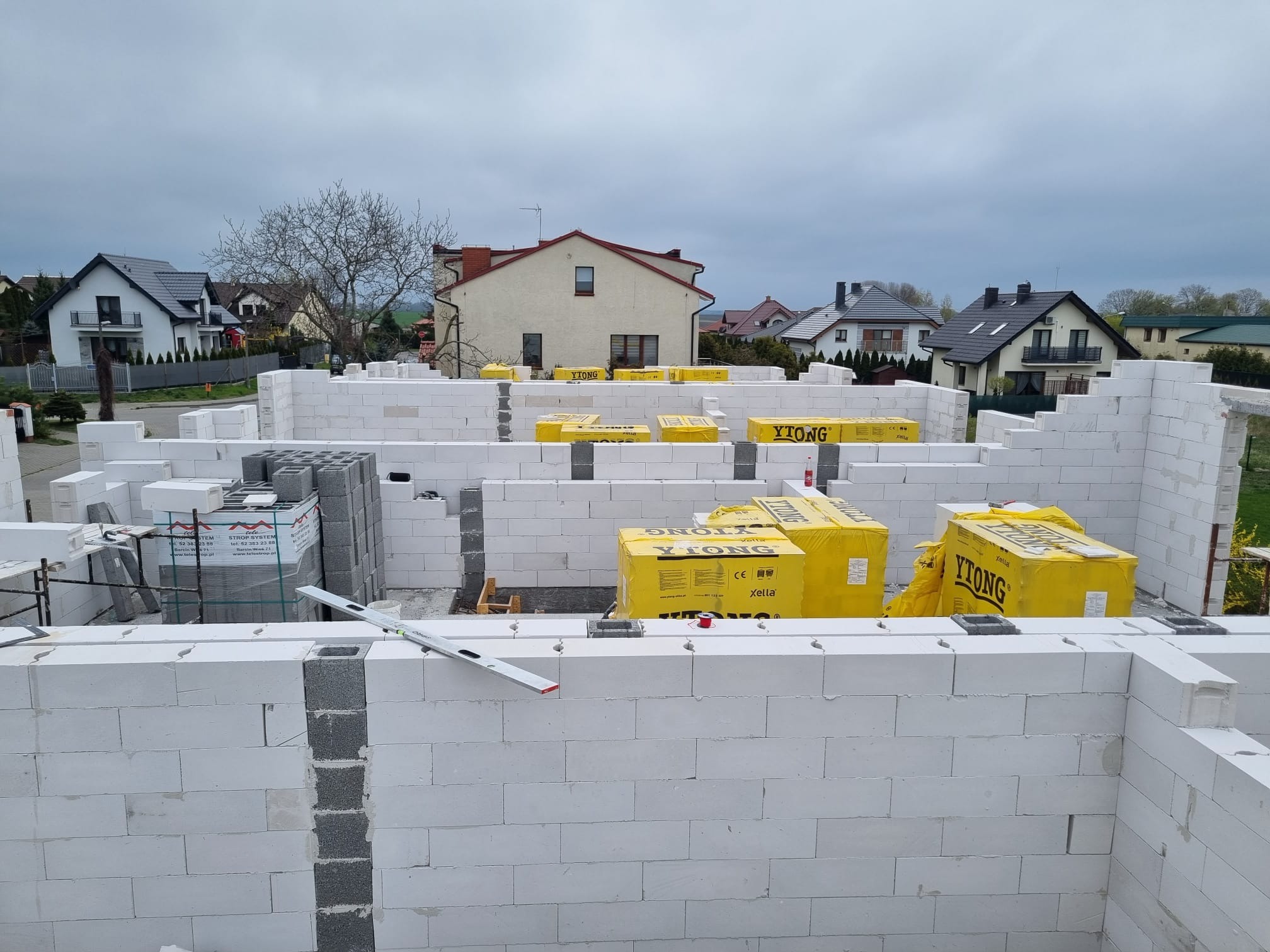 Budowa ścian domu z bloczków Ytong, widoczne palety z materiałem budowlanym oraz poziomica na murze, w tle zabudowa jednorodzinna.