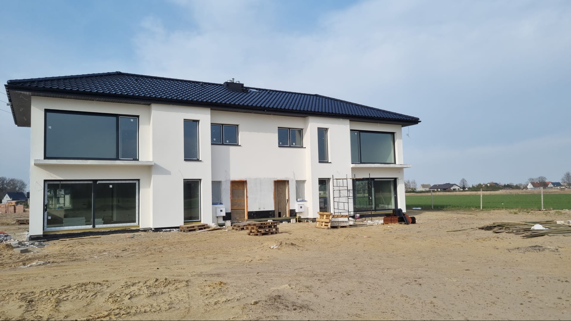 Bliźniak w trakcie budowy, z białymi ścianami, dużymi oknami i czarnym dachem, widoczne rusztowania i materiały budowlane.