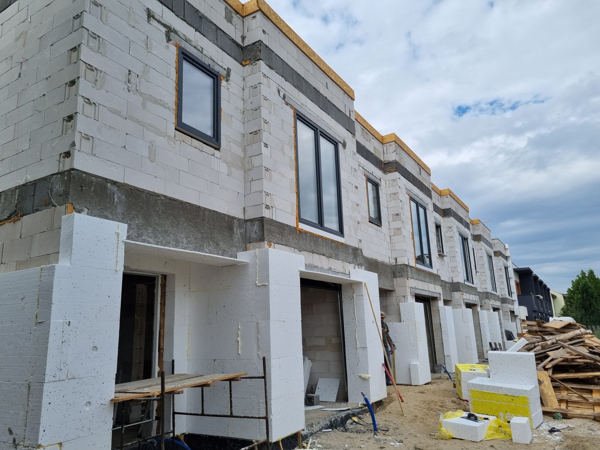 Szeregówka w stanie surowym otwartym z częściowo zamontowaną stolarką okienną i przygotowanymi otworami garażowymi obłożonymi styropianem, widoczne materiały budowlane na terenie budowy.