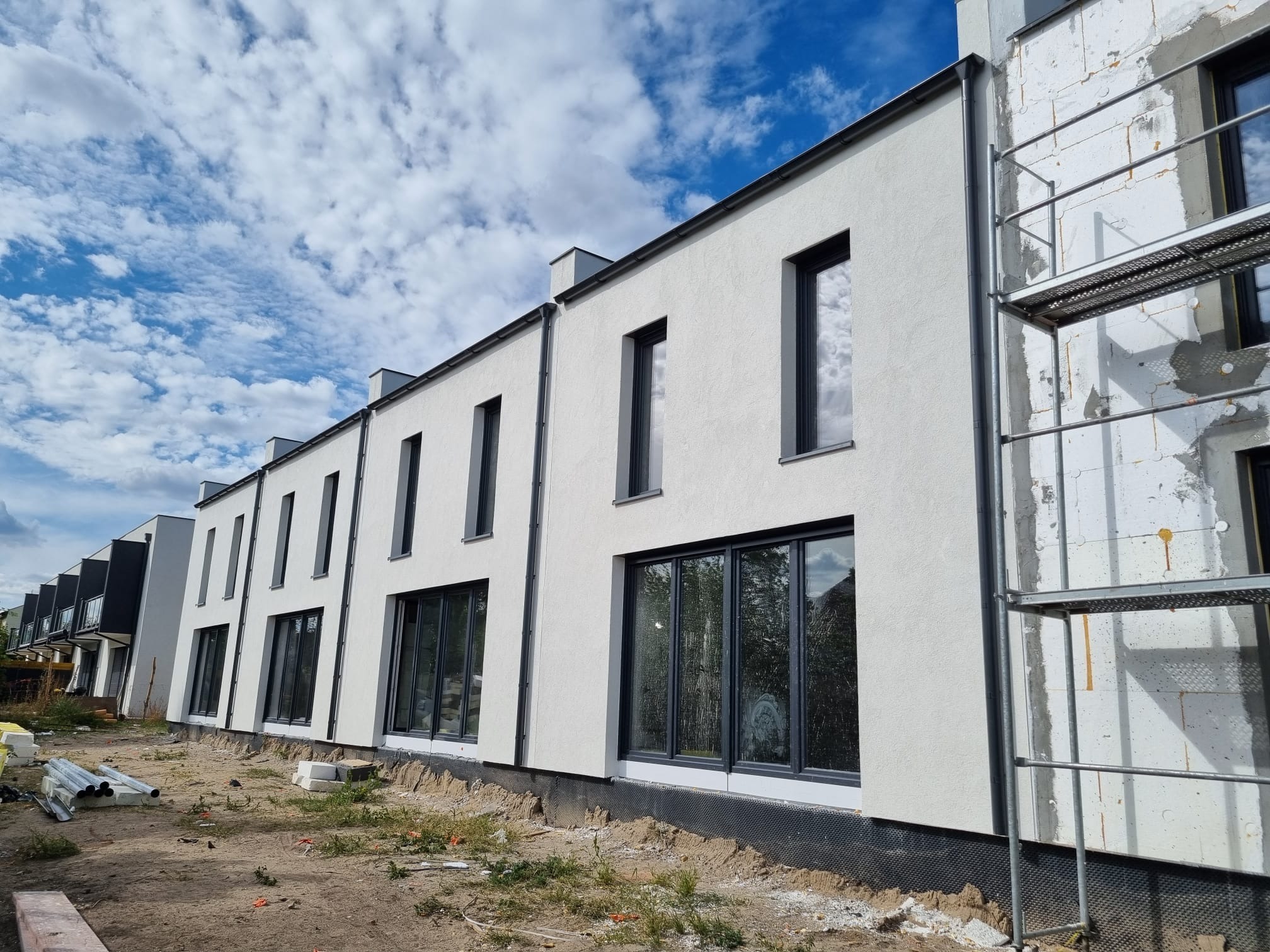 Rząd szeregowych domów jednorodzinnych w trakcie wykańczania elewacji, jeden z domów z widocznym rusztowaniem i nieotynkowaną ścianą, pozostałe z białą elewacją i ciemnymi oknami, błękitne niebo...