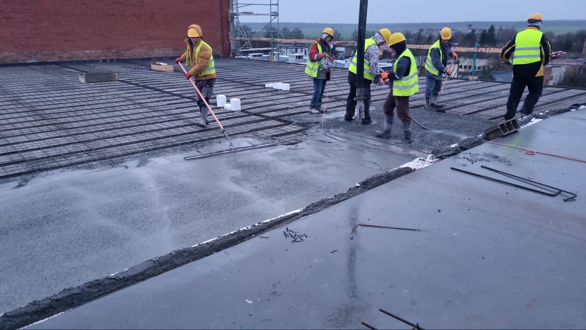 Ekipa budowlana w żółtych kamizelkach i kaskach, w trakcie wylewania i rozprowadzania betonu na zbrojonej powierzchni dachu budynku w szary, pochmurny dzień.