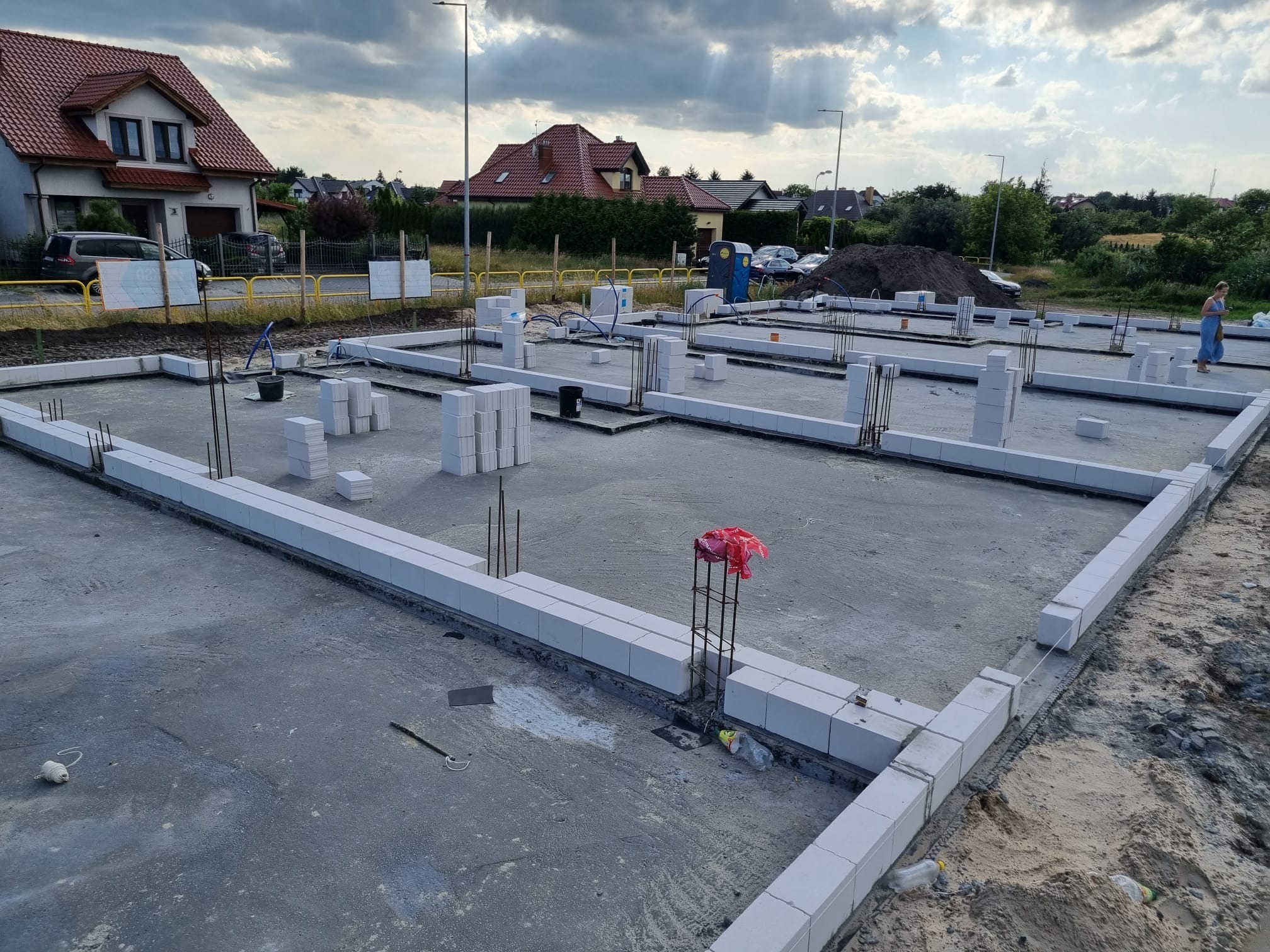 Zalany fundament z bloczków betonowych, wystające pręty zbrojeniowe, widoczne zarysy przyszłych ścian, w tle zabudowa jednorodzinna i pochmurne niebo.