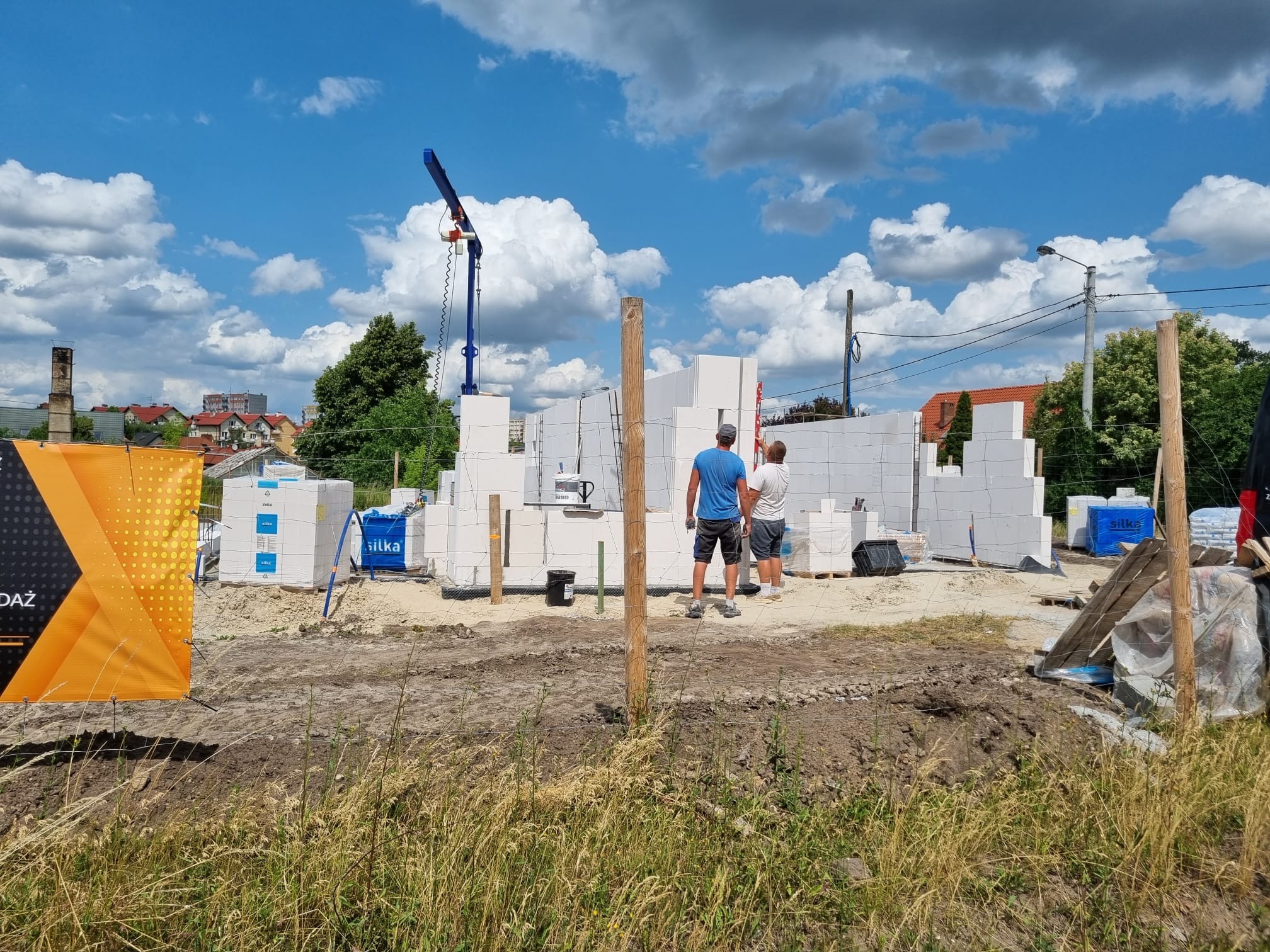 Budowa domu z białych bloczków silikatowych, widoczny dźwig, dwóch pracowników przy ścianie, palety z materiałem budowlanym i baner reklamowy w tle błękitne niebo z chmurami.