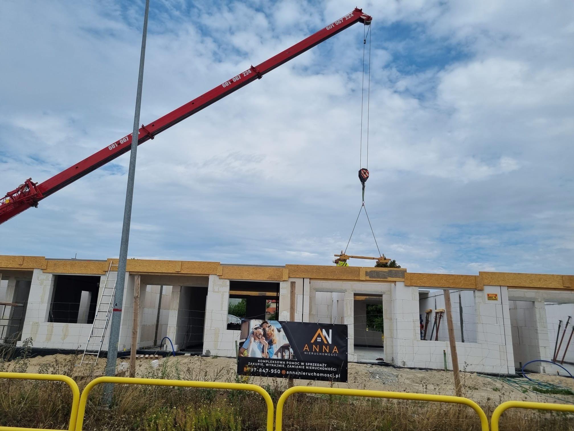 Budowa domu z białych bloczków: czerwony dźwig podnosi element konstrukcyjny, widoczny baner reklamowy 'ANNA Nieruchomości' oraz drabina oparta o ścianę.