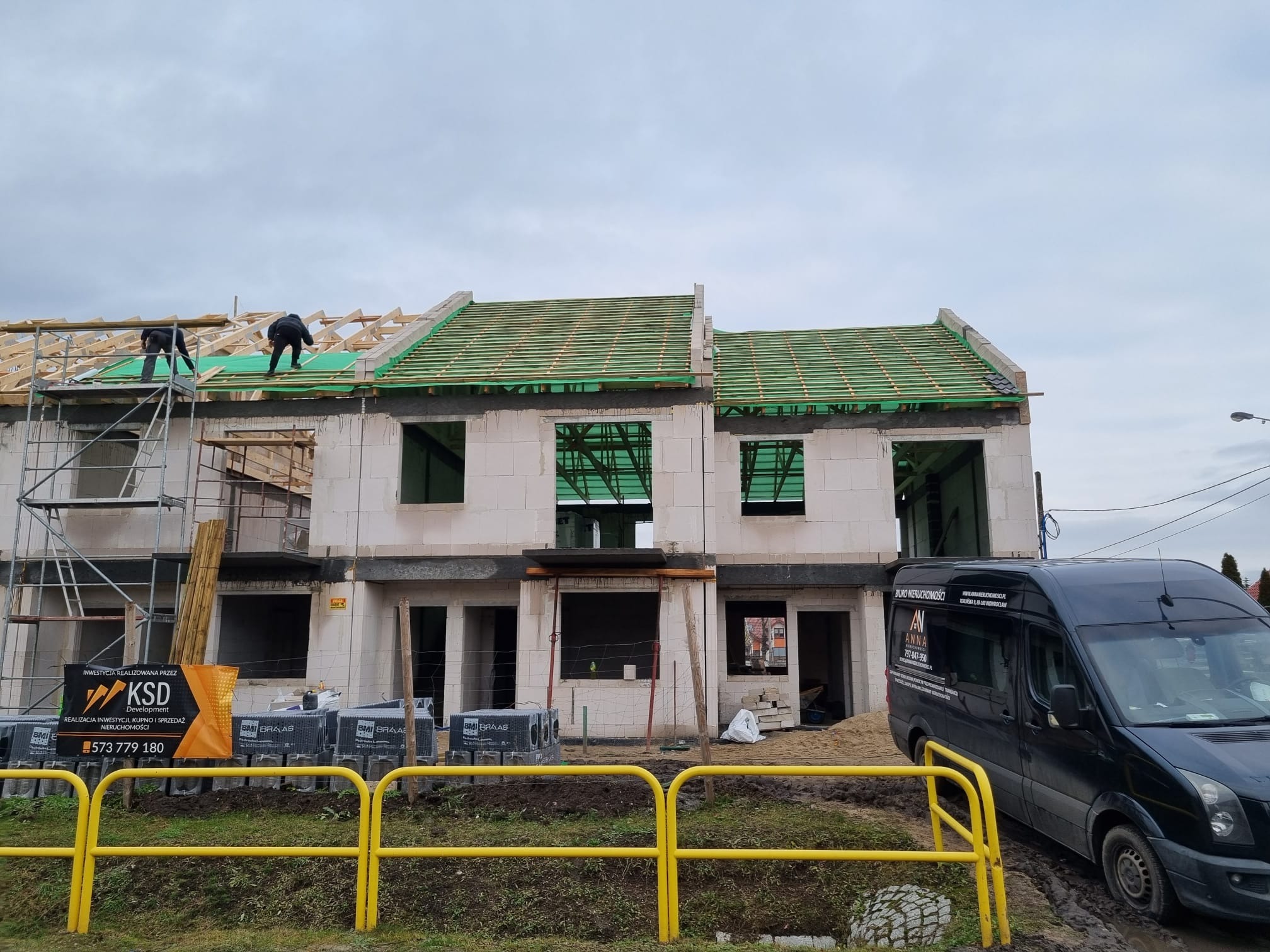 Budowa bliźniaka w stanie surowym otwartym, widoczne rusztowania, ekipa montująca konstrukcję dachu oraz zaparkowany bus firmy budowlanej.