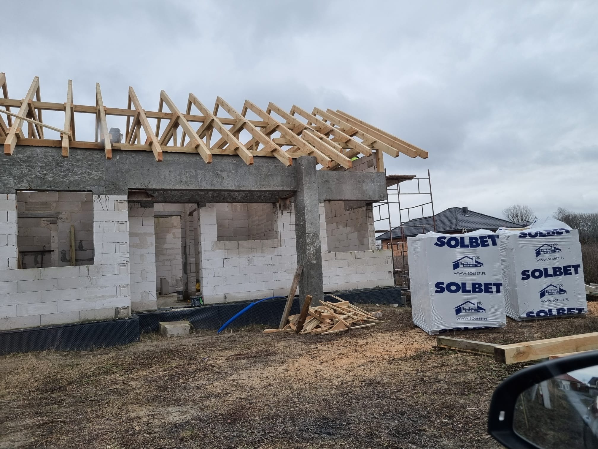 Surowy stan domu w trakcie budowy: ściany z białych bloczków, drewniana konstrukcja dachu, paczki materiałów budowlanych Solbet z logo producenta na działce.