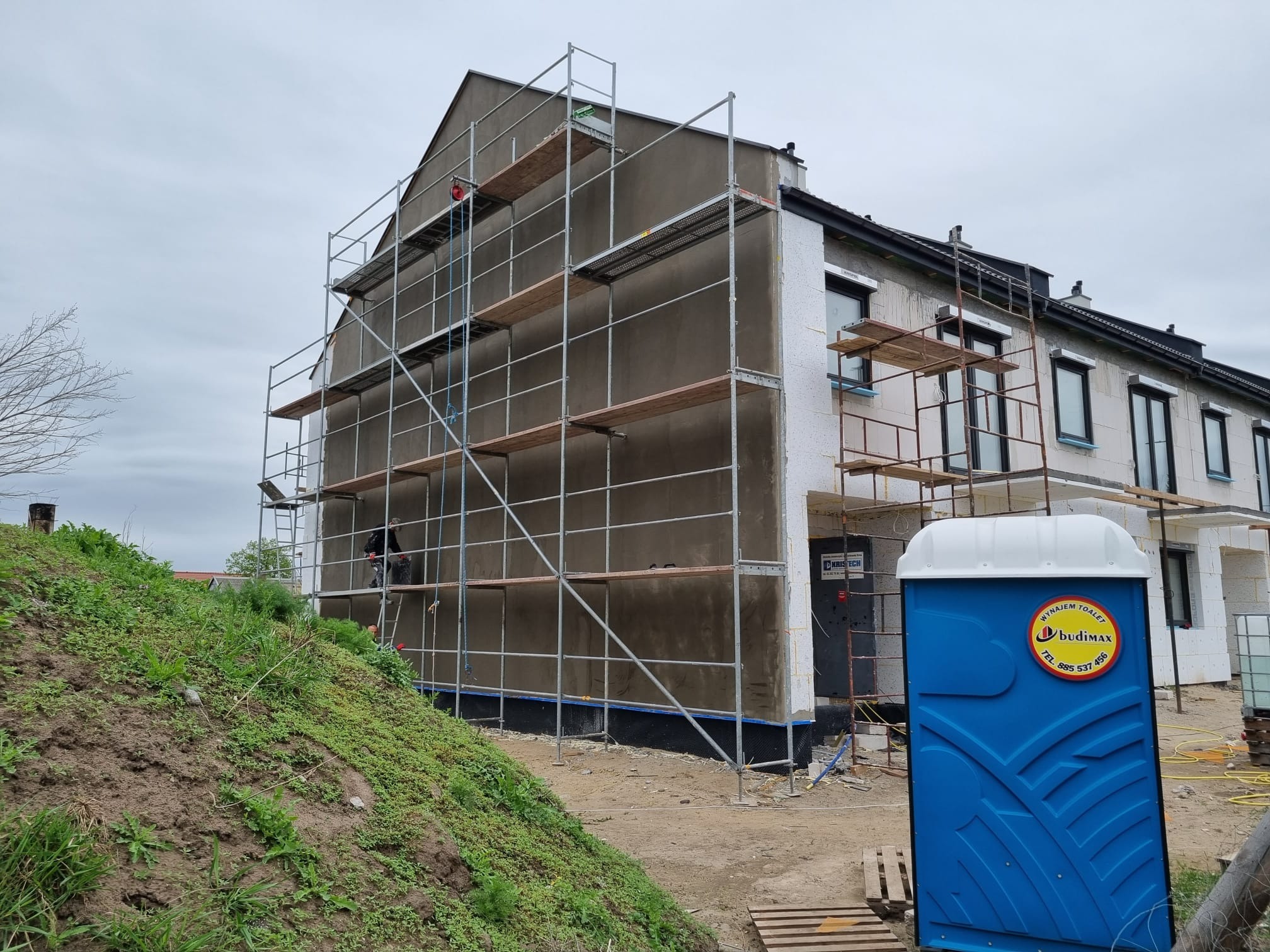 Budynek w trakcie prac elewacyjnych, widoczne rusztowania, częściowo ocieplona ściana i przenośna toaleta na pierwszym planie.