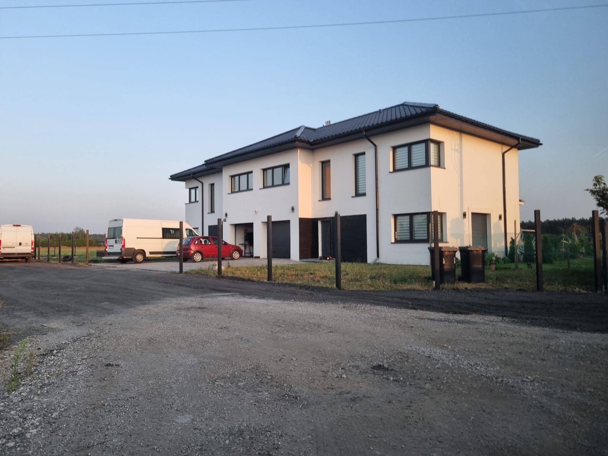 Nowoczesny, dwukondygnacyjny dom w zabudowie bliźniaczej z białą elewacją i ciemnymi oknami w Inowrocławiu, widoczny z perspektywy podjazdu z dwoma samochodami dostawczymi i osobowym.