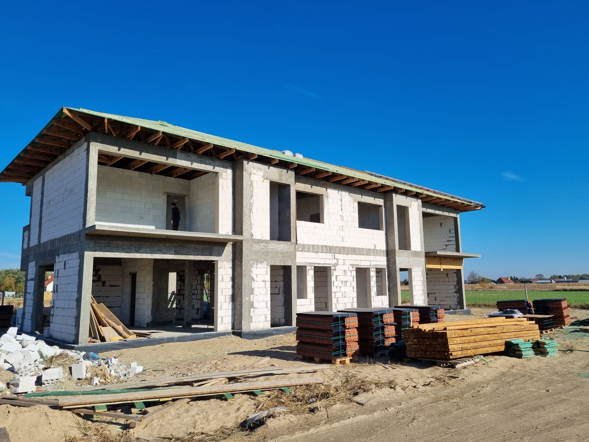 Surowy stan dwukondygnacyjnego domu z białych bloczków, widoczna konstrukcja dachu z zieloną membraną, stosy dachówek i drewna na placu budowy, błękitne niebo w tle.