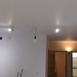 M&G Grzegorz Koziński - Wnętrze świeżo pomalowanego pokoju z zamontowanymi reflektorami sufitowymi i wiszącą żarówką na kablu, widoczne ślady po gniazdkach elektrycznych.