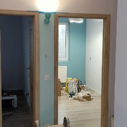 M&G Grzegorz Koziński - Dwa otwarte, drewniane drzwi wewnętrzne w nowo wykończonym pokoju z pomalowanymi na biało ścianami i turkusowym akcentem na jednej ze ścian; widoczne worki z materiałami budowlanymi w tle.