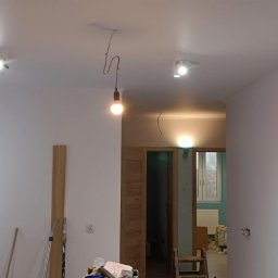 M&G Grzegorz Koziński - Wnętrze w trakcie prac wykończeniowych: świeżo pomalowane ściany na biało, widoczne punkty świetlne na suficie, wisząca żarówka na kablu, przygotowane narzędzia i materiały budowlane na podłodze.