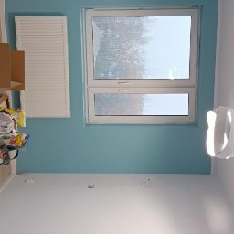 M&G Grzegorz Koziński - Świeżo pomalowane na turkusowo górne partie ścian w pokoju z białymi ścianami poniżej, grzejnikiem i oknem z widokiem na drzewa.