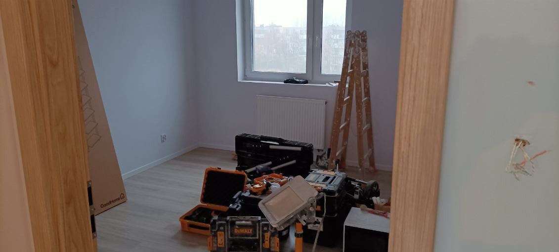 Pokój w trakcie prac wykończeniowych, widoczne narzędzia DeWalt, drabina, skrzynki narzędziowe, ściany pomalowane na jasny kolor, widok przez futrynę drzwi.