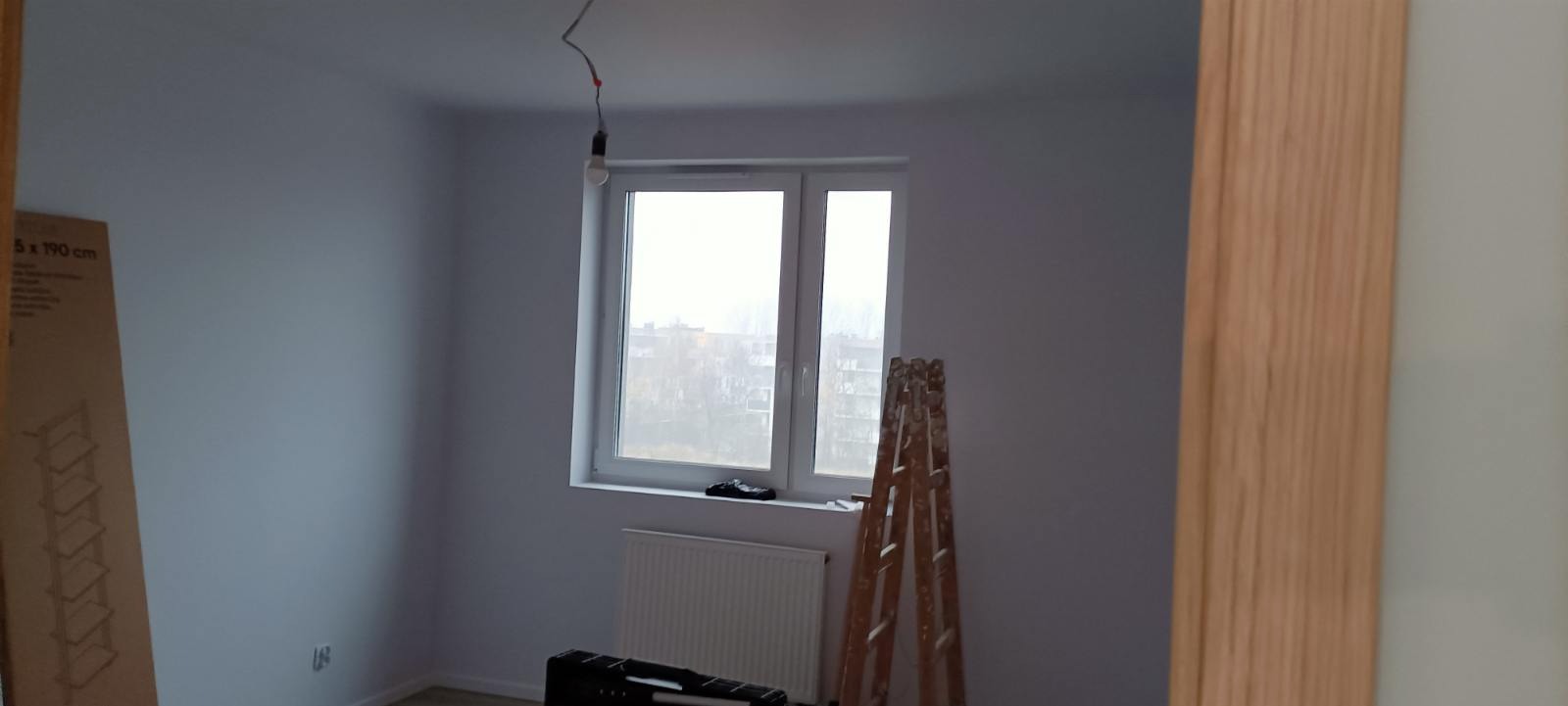 Pokój po malowaniu na szaro, z drewnianą drabiną opartą o ścianę przy oknie, widoczne kartonowe opakowanie regału i wisząca żarówka.