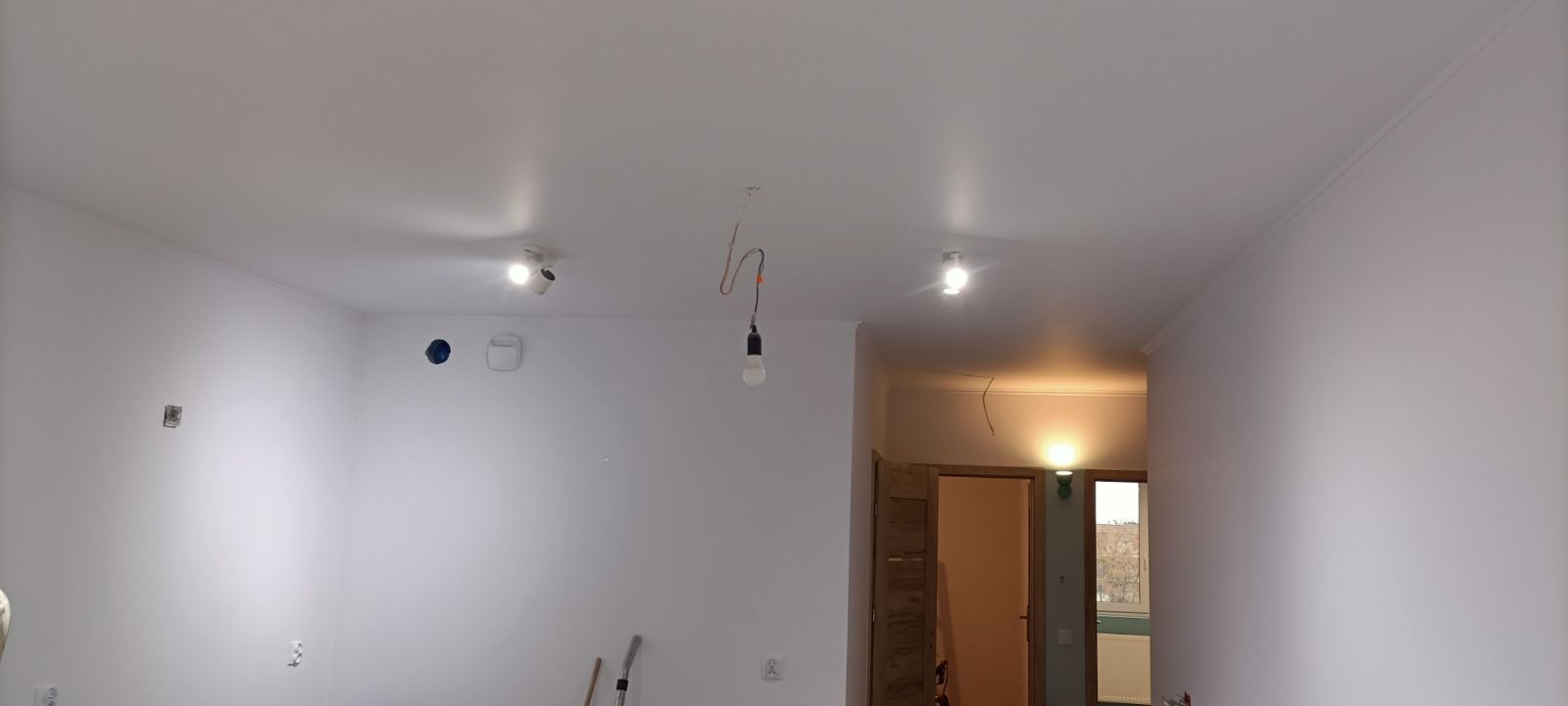 Wnętrze świeżo pomalowanego pokoju z zamontowanymi reflektorami sufitowymi i wiszącą żarówką na kablu, widoczne ślady po gniazdkach elektrycznych.