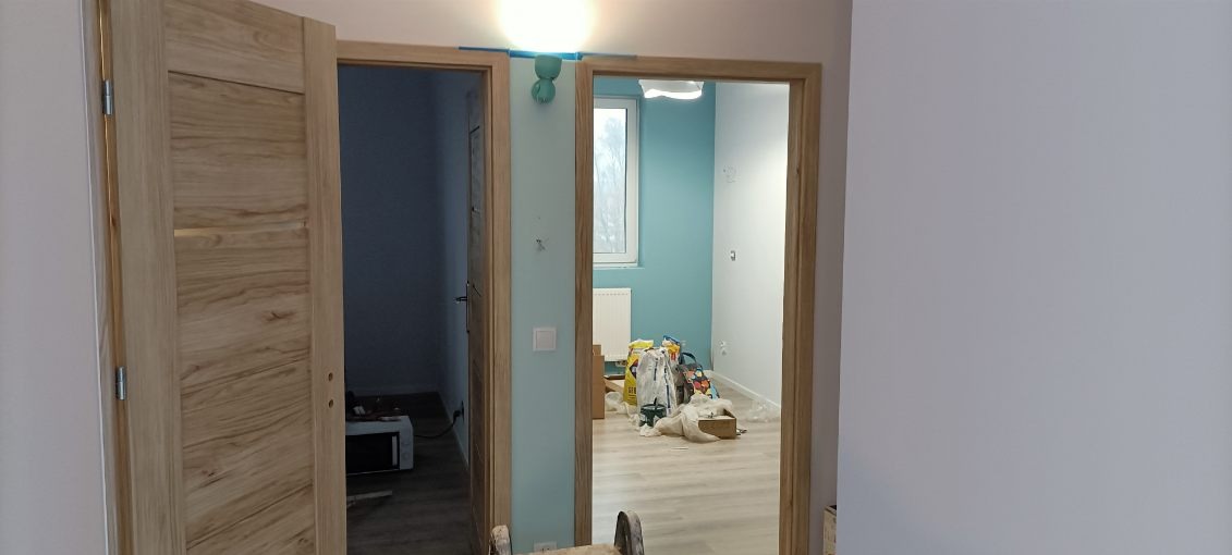 Dwa otwarte, drewniane drzwi wewnętrzne w nowo wykończonym pokoju z pomalowanymi na biało ścianami i turkusowym akcentem na jednej ze ścian; widoczne worki z materiałami budowlanymi w tle.