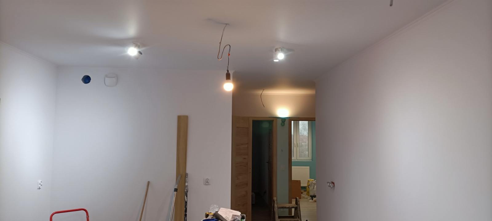 Wnętrze w trakcie prac wykończeniowych: świeżo pomalowane ściany na biało, widoczne punkty świetlne na suficie, wisząca żarówka na kablu, przygotowane narzędzia i materiały budowlane na podłodze.