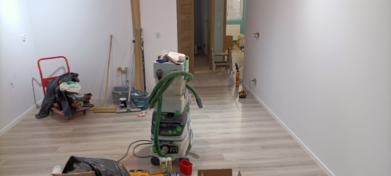 Wnętrze w trakcie remontu: świeżo pomalowane białe ściany, panele podłogowe, narzędzia budowlane, odkurzacz przemysłowy, wózek transportowy z narzędziami, widoczne gniazdka elektryczne.