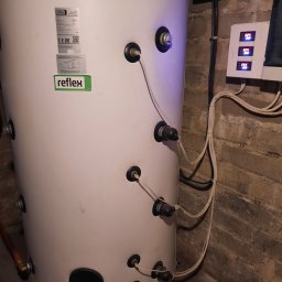 OZE Level Sławomir Dybcio - Biały zbiornik buforowy Reflex z podłączonymi przewodami w kotłowni w Warce, obok widoczny panel sterowania z wyświetlanymi temperaturami.