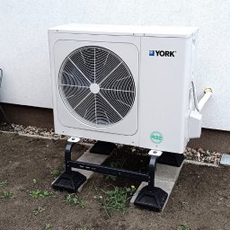 OZE Level Sławomir Dybcio - Biała pompa ciepła marki York, zamontowana na czarnej metalowej podstawie i betonowych płytach, widoczna na tle białej ściany budynku i ziemi z kamykami.