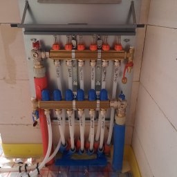 OZE Level Sławomir Dybcio - Rozdzielacz ogrzewania podłogowego zamontowany na ścianie z pustaków, widoczne rury doprowadzające i odprowadzające wodę, oraz maty grzewcze na podłodze w tle.