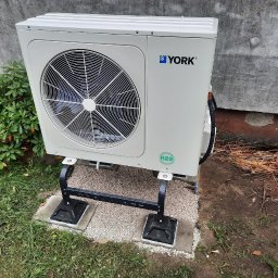 OZE Level Sławomir Dybcio - Zewnętrzna jednostka pompy ciepła marki York zamontowana na czarnym metalowym stelażu, posadowiona na betonowych płytach i żwirze, z zieloną roślinnością i fragmentem ściany w tle.