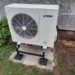 Zewnętrzna jednostka pompy ciepła marki York zamontowana na czarnym metalowym stelażu, posadowiona na betonowych płytach i żwirze, z zieloną roślinnością i fragmentem ściany w tle.