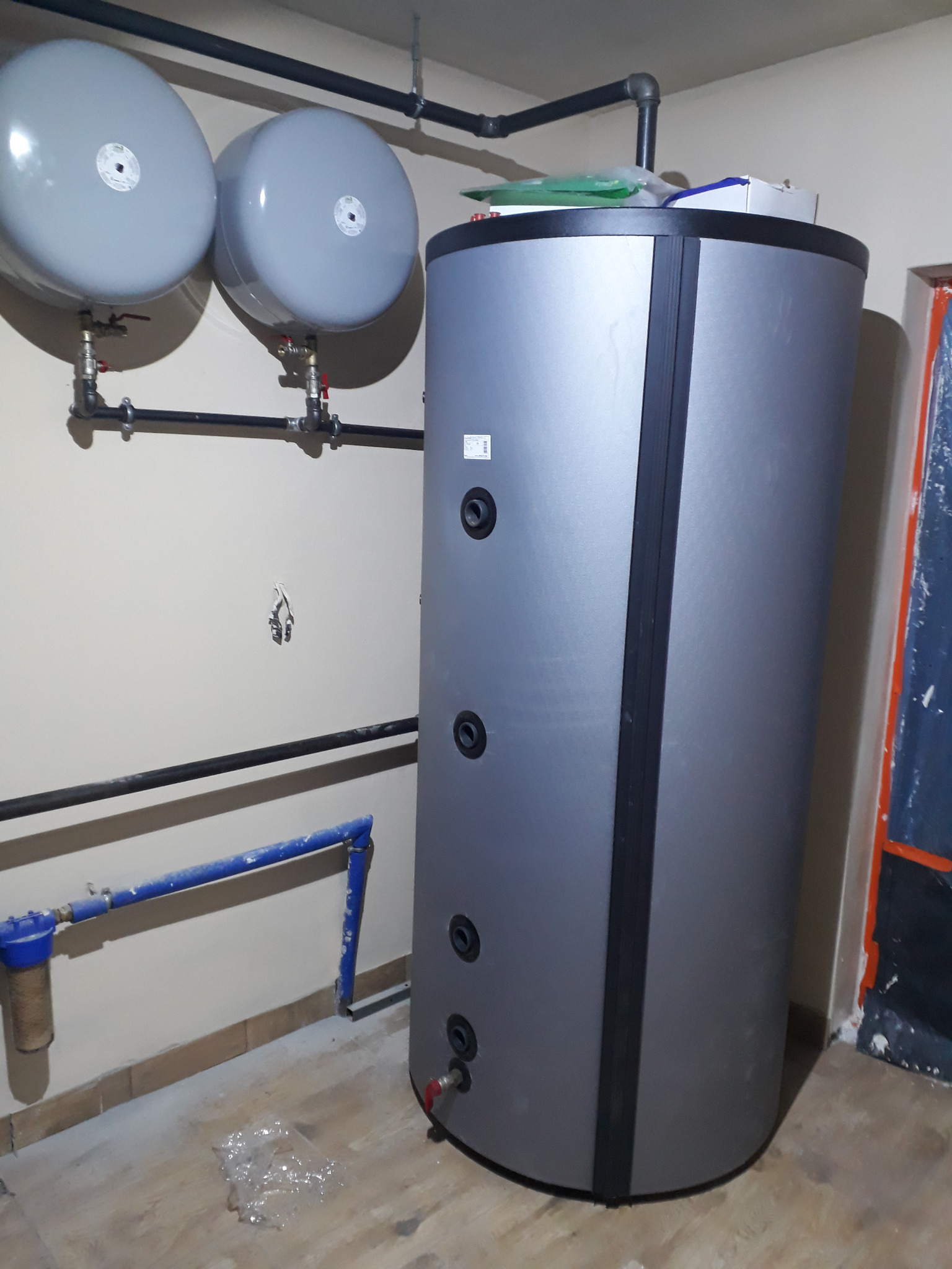 Szary, cylindryczny zbiornik buforowy z podłączonymi rurami, widoczne dwa mniejsze zbiorniki wyrównawcze na ścianie, filtr na rurze doprowadzającej wodę, podłoga w trakcie prac wykończeniowych.
