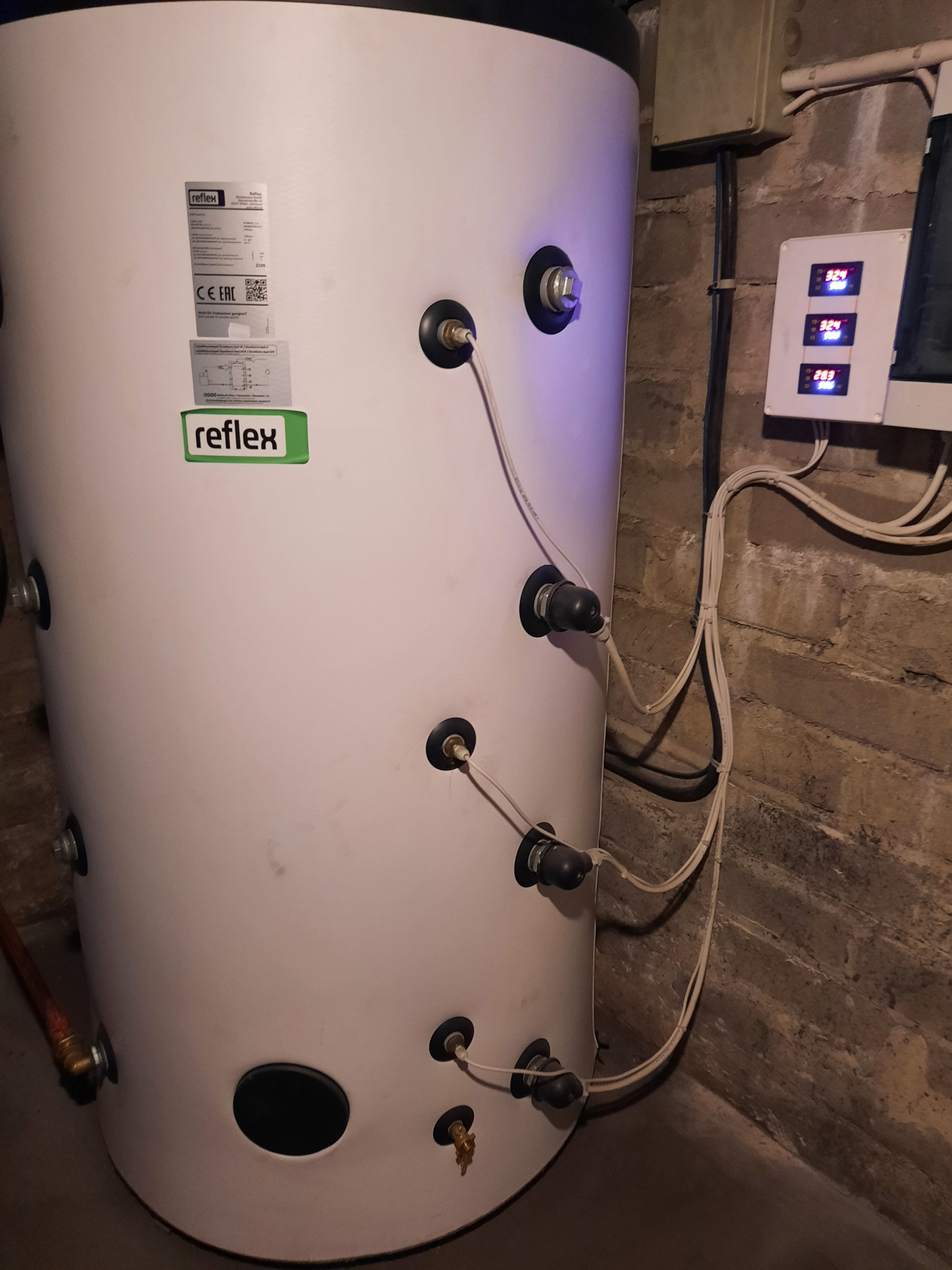Biały zbiornik buforowy Reflex z podłączonymi przewodami w kotłowni w Warce, obok widoczny panel sterowania z wyświetlanymi temperaturami.