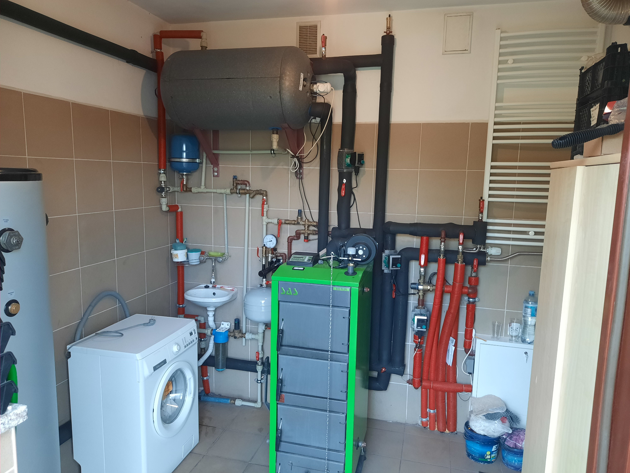 Kotłownia z piecem na paliwo stałe, zasobnikiem ciepłej wody użytkowej, pralką i umywalką, instalacja grzewcza z rurami w kolorze czerwonym i czarnym, widoczne elementy instalacji hydraulicznej.
