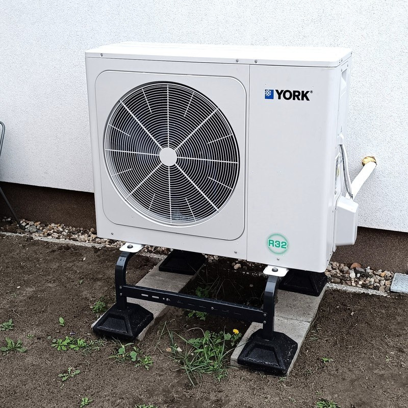 Biała pompa ciepła marki York, zamontowana na czarnej metalowej podstawie i betonowych płytach, widoczna na tle białej ściany budynku i ziemi z kamykami.