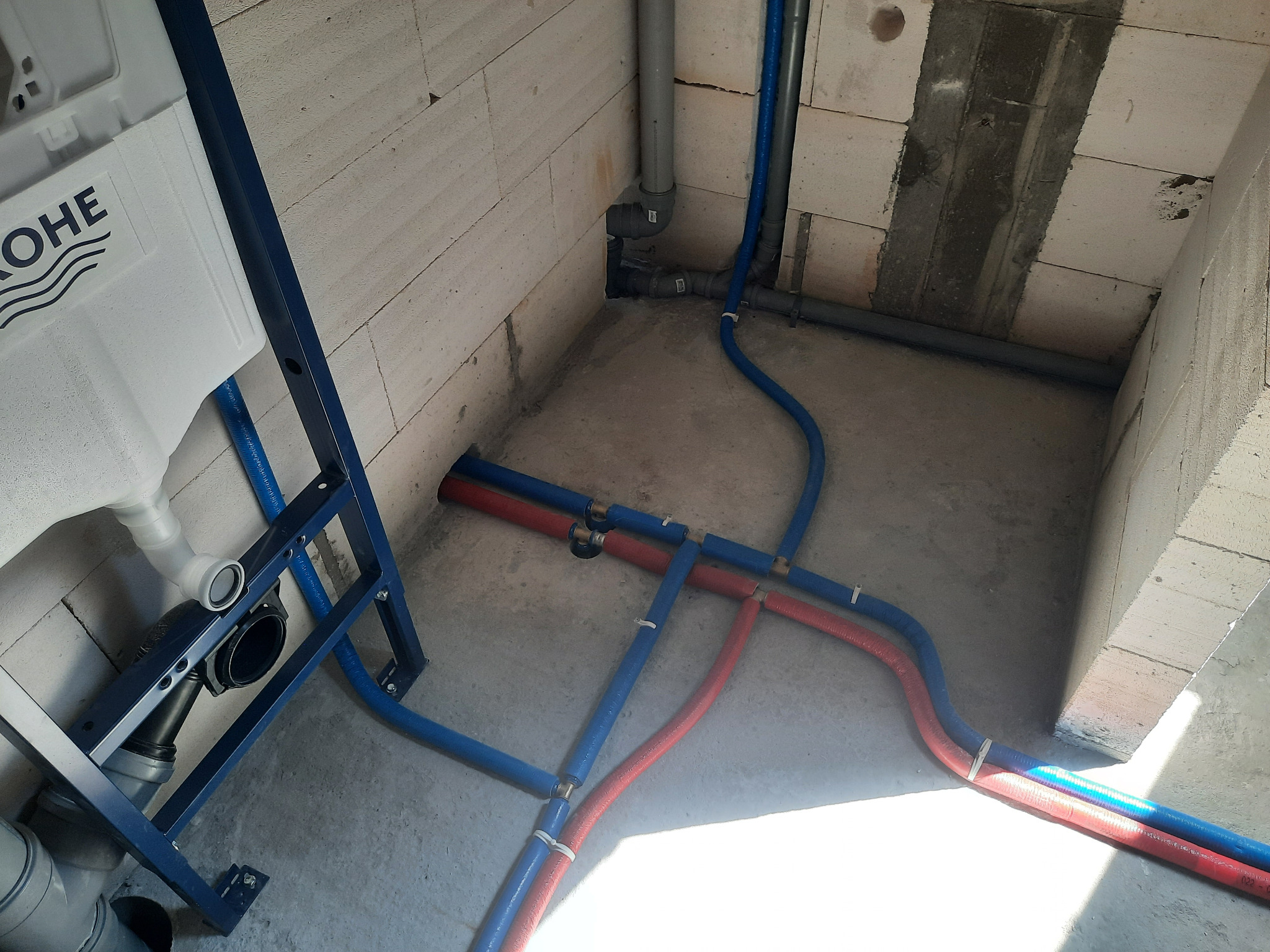 Szare rury kanalizacyjne i niebiesko-czerwone rury doprowadzające wodę, zamocowane do ściany z pustaków, z widocznym stelażem podtynkowym WC marki Grohe.