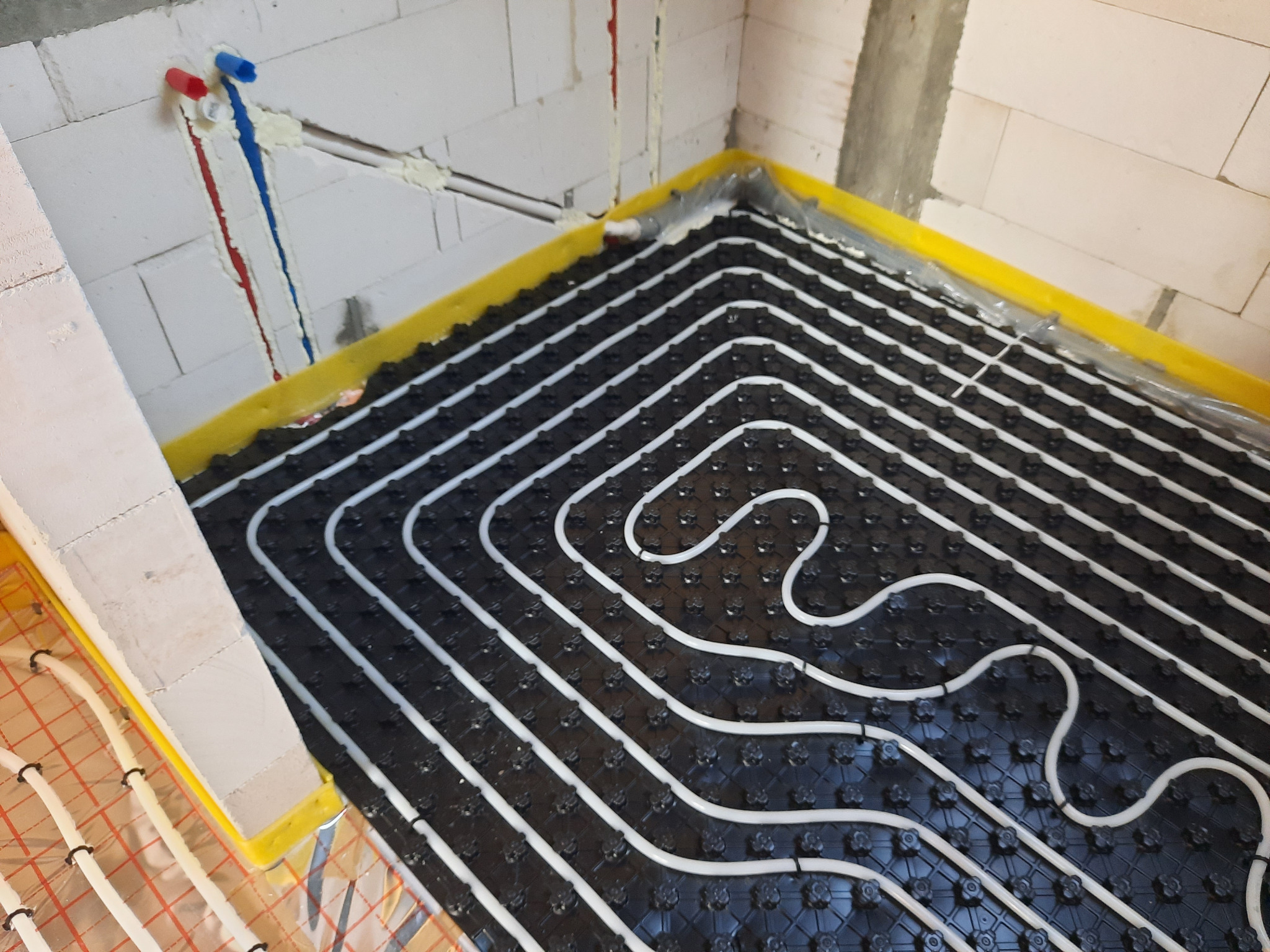 Rozłożona instalacja ogrzewania podłogowego z białymi rurkami na czarnej macie izolacyjnej, widoczne pionowe rury doprowadzające wodę z oznaczeniami kolorami ciepłej i zimnej wody przy ścianie...