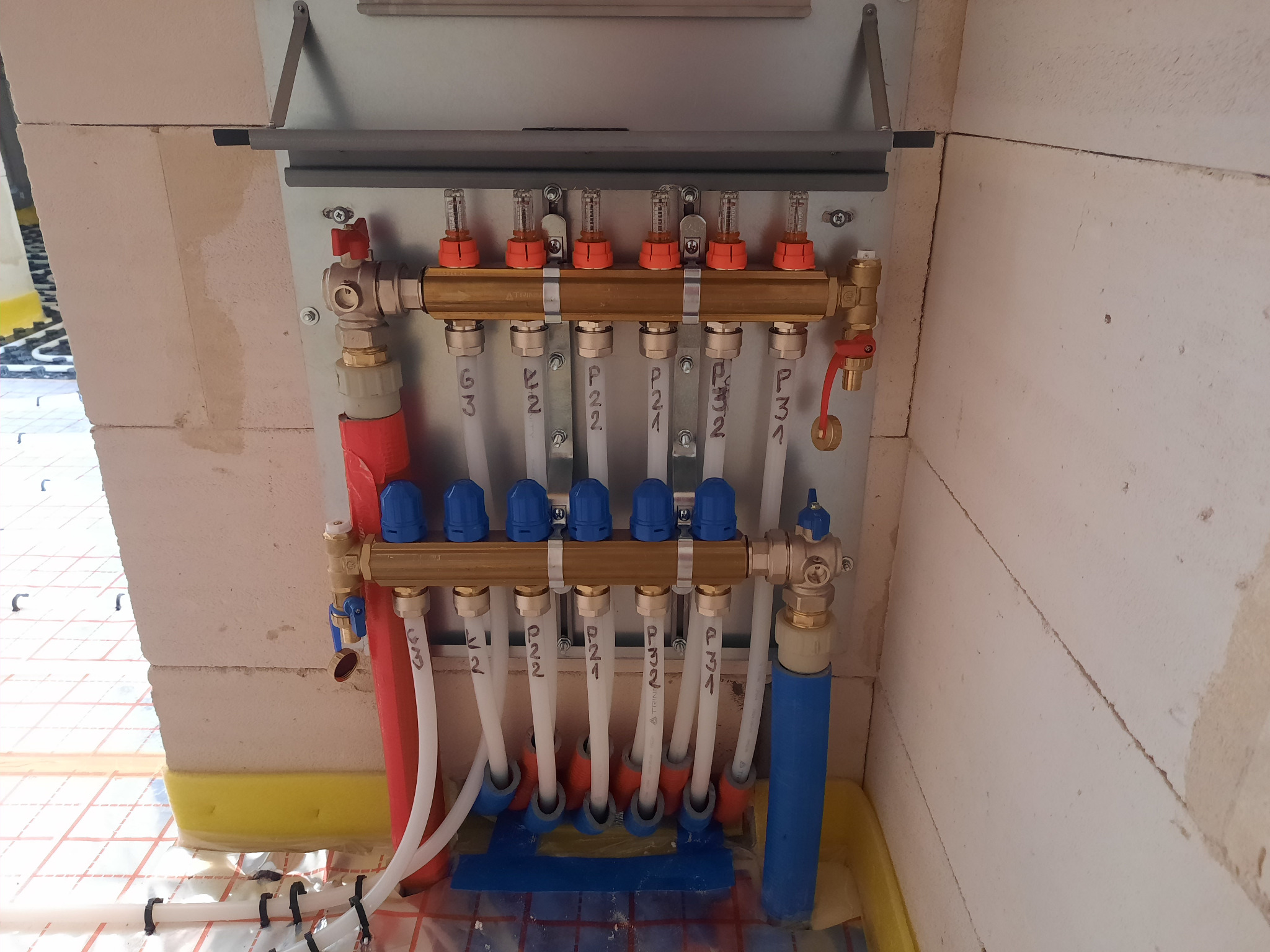 Rozdzielacz ogrzewania podłogowego zamontowany na ścianie z pustaków, widoczne rury doprowadzające i odprowadzające wodę, oraz maty grzewcze na podłodze w tle.