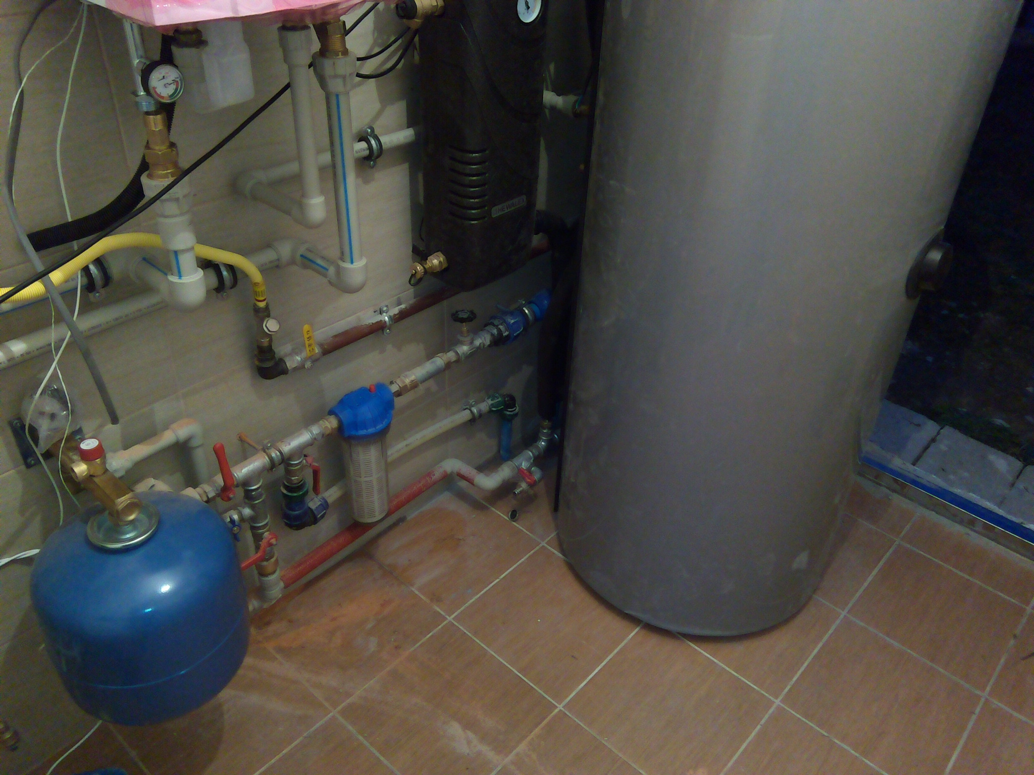 Instalacja kotła z filtrem wody, zbiornikiem wyrównawczym i kompletem rur w kotłowni wyłożonej brązowymi płytkami podłogowymi.