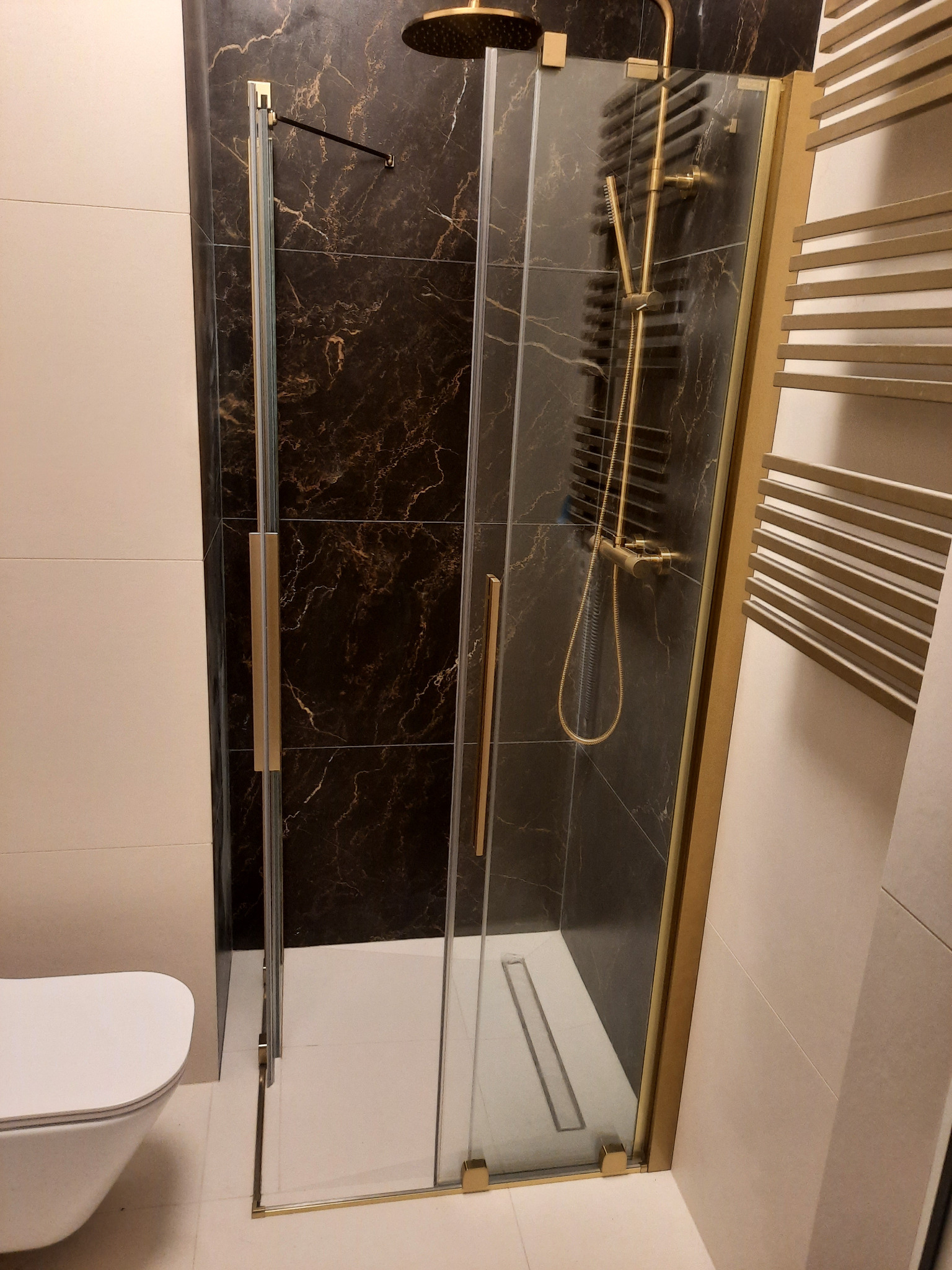 Kabina prysznicowa z czarnymi marmurowymi płytkami, złotymi okuciami i grzejnikiem drabinkowym w łazience z jasnymi płytkami.