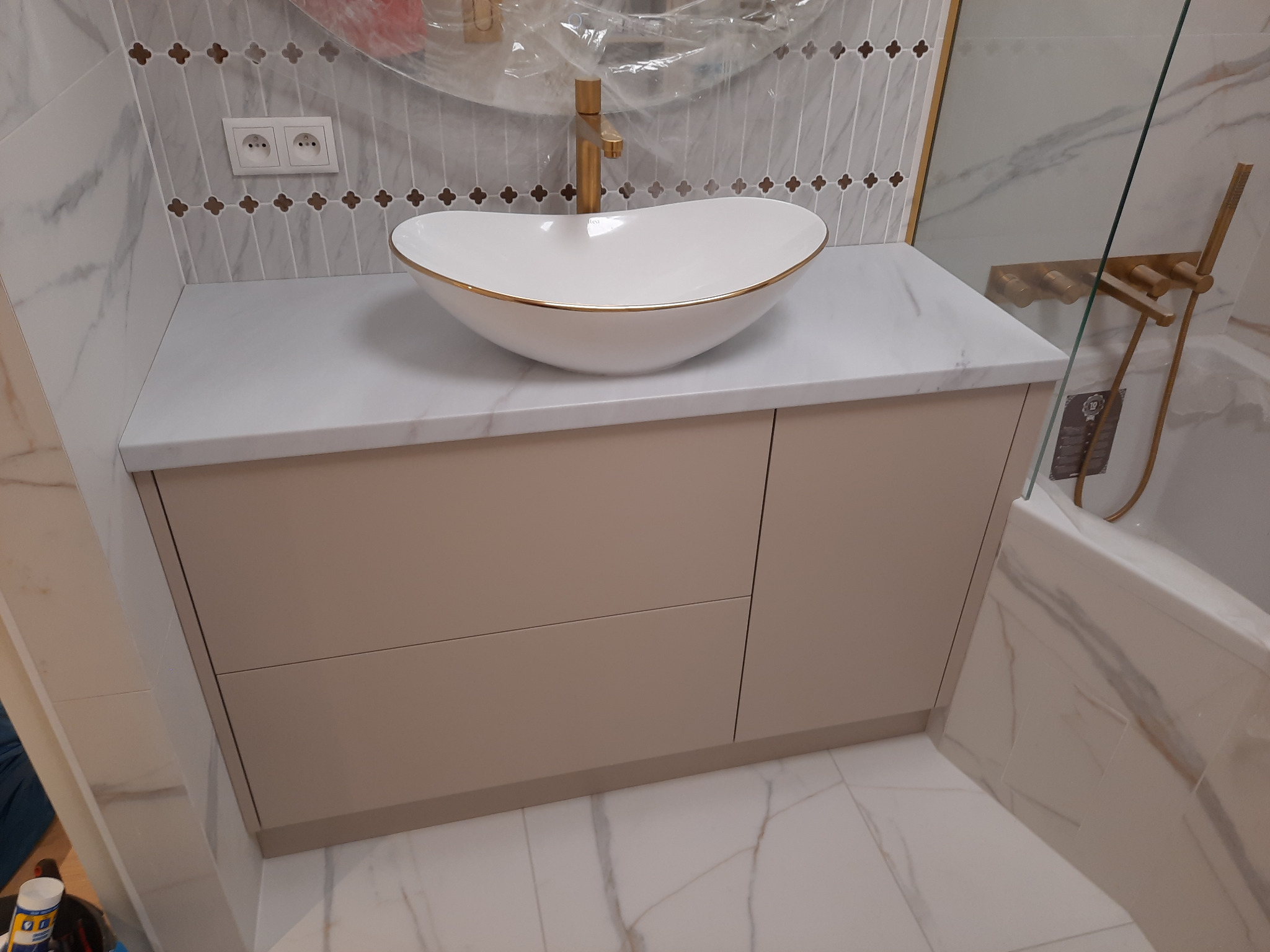 Umywalka nablatowa w kształcie łzy z złotym rantem i baterią w kolorze złota, zamontowana na beżowej szafce łazienkowej z szufladami, na tle marmurowych płytek i fragmentu kabiny prysznicowej...