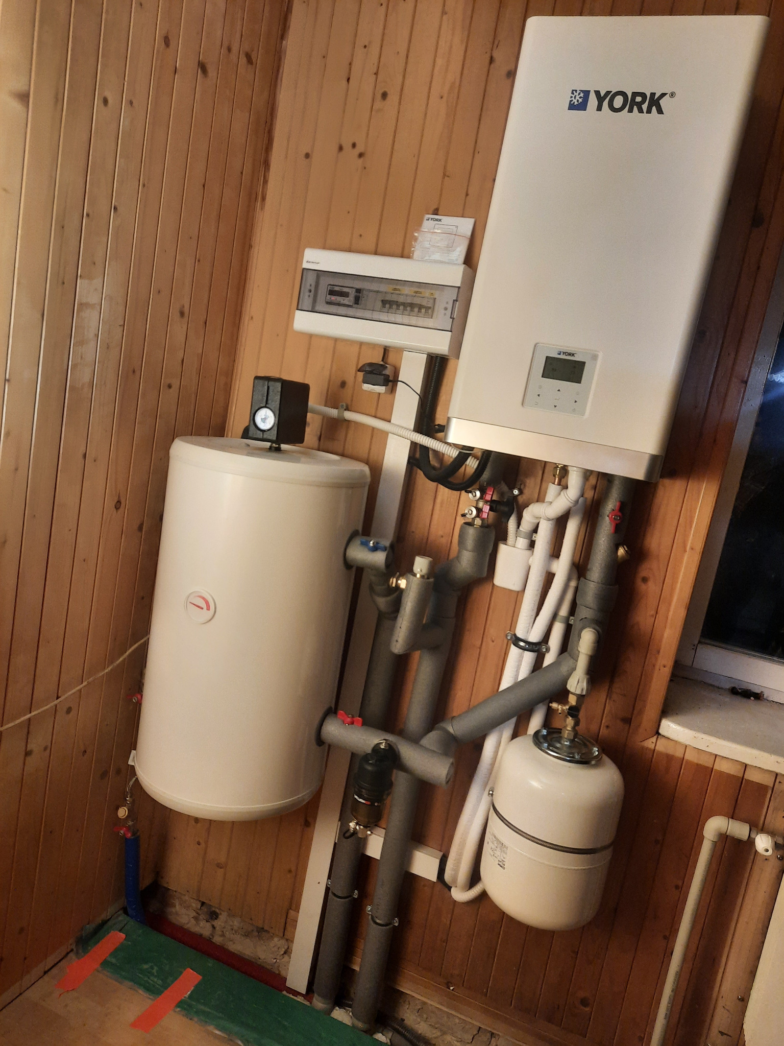 Biały piec gazowy marki York, biały zasobnik ciepłej wody i białe naczynie przeponowe zamontowane na ścianie obitej boazerią, widoczne szare i białe rury instalacyjne.