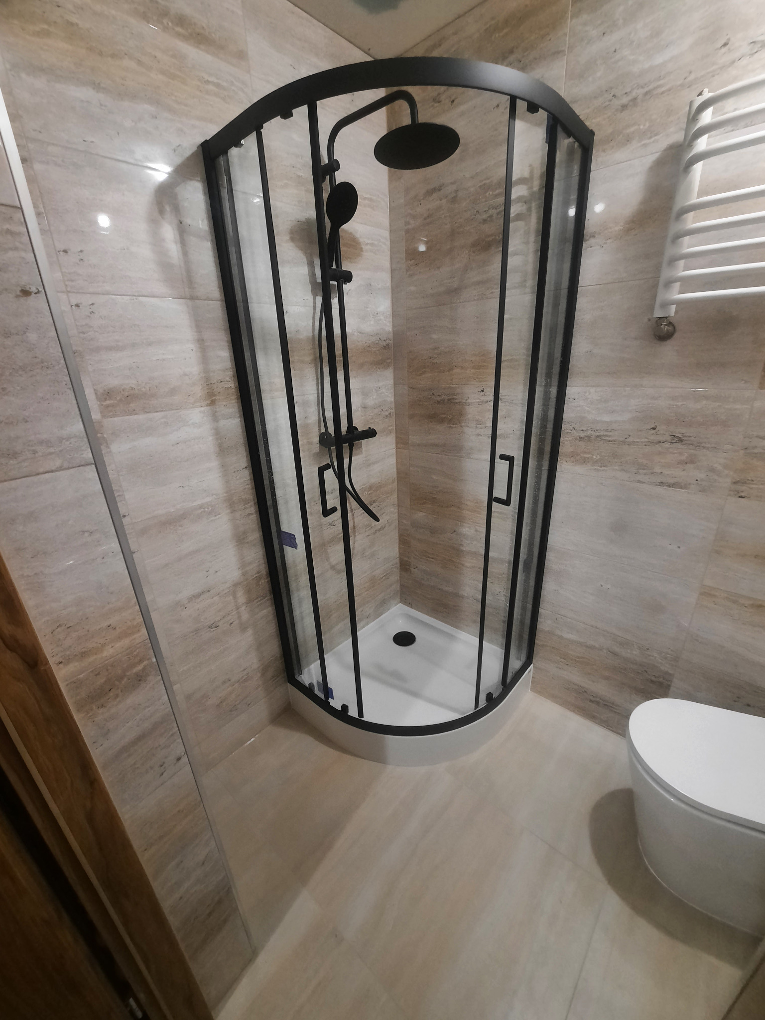 Nowoczesna kabina prysznicowa z czarnymi ramami i deszczownicą w łazience wykończonej beżowymi płytkami imitującymi marmur, widoczny również biały grzejnik drabinkowy i toaleta wisząca.