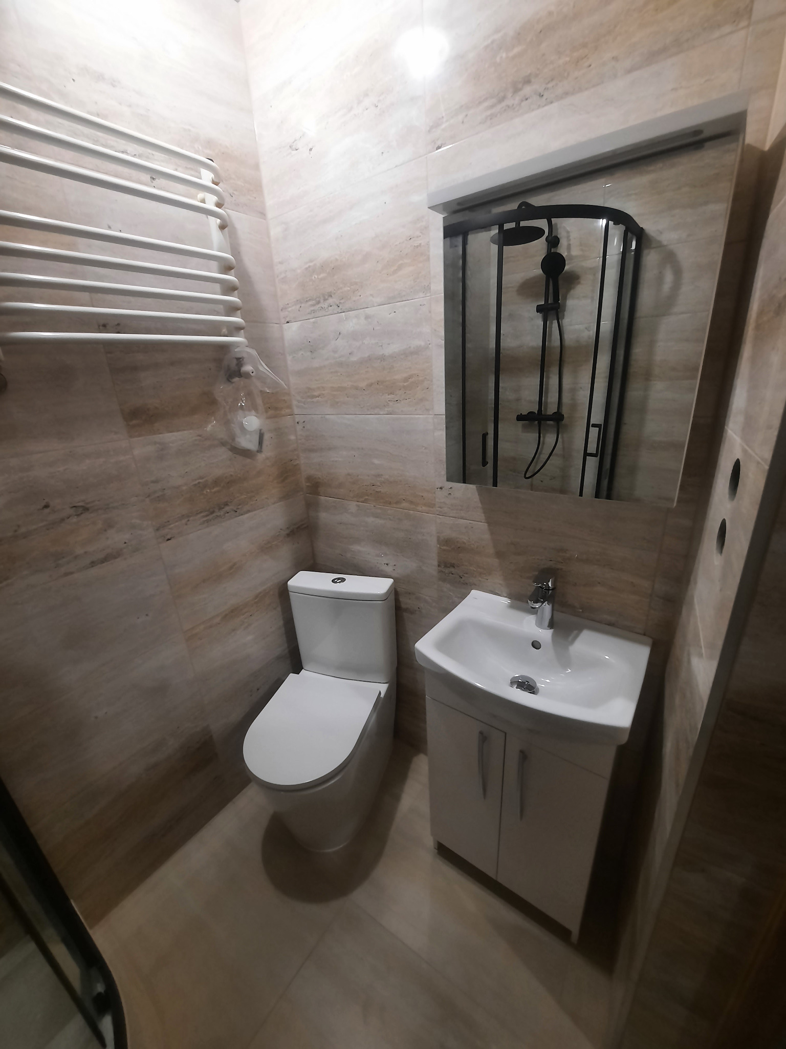 Nowoczesna łazienka z beżowymi płytkami imitującymi marmur, wyposażona w białą toaletę, umywalkę z szafką i czarną kabinę prysznicową z deszczownicą, widoczny również grzejnik drabinkowy.