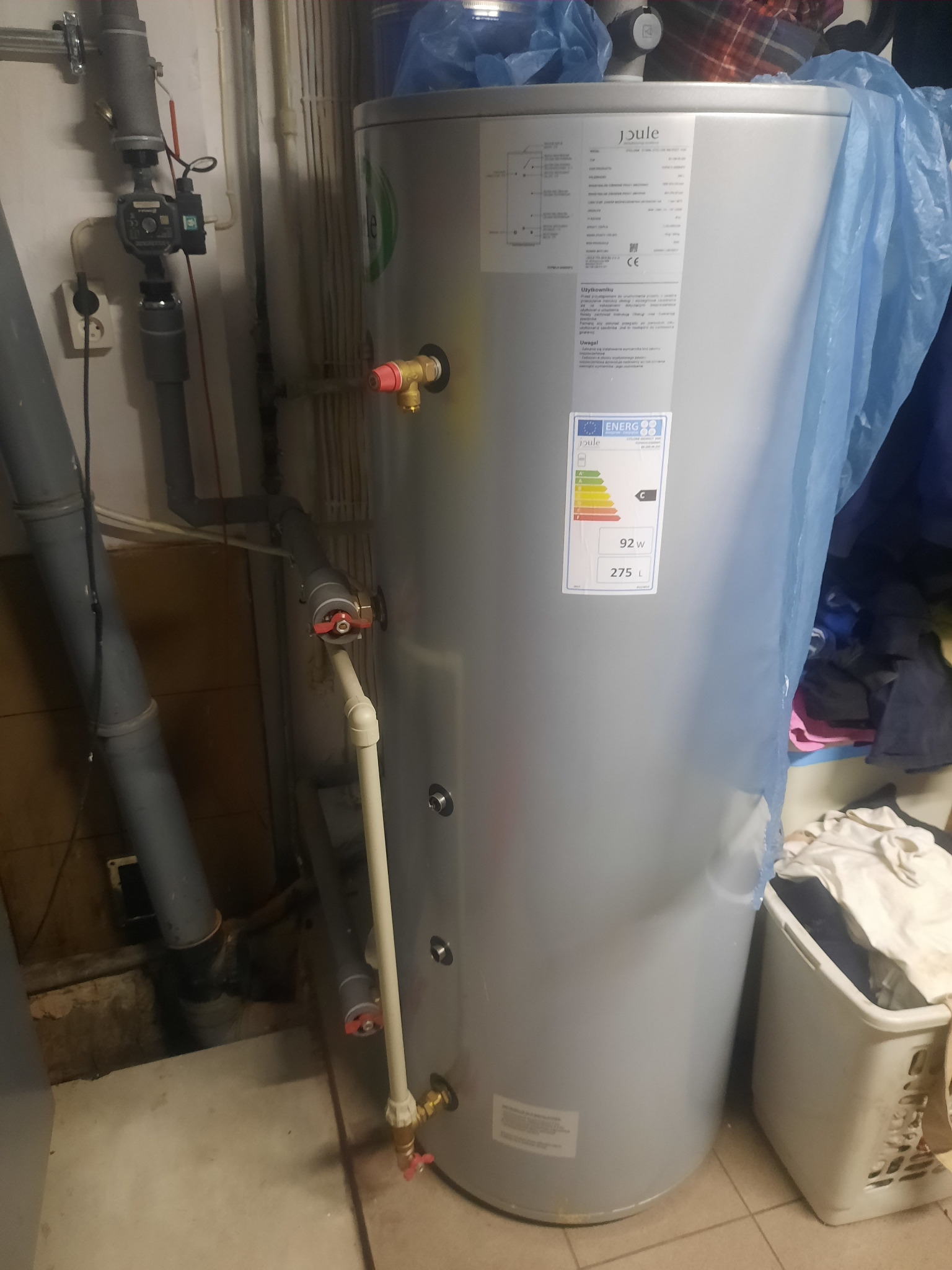 Szary, pionowy zbiornik na wodę marki Joule z etykietą energetyczną, widoczne rury z zaworami, kosz na pranie w tle