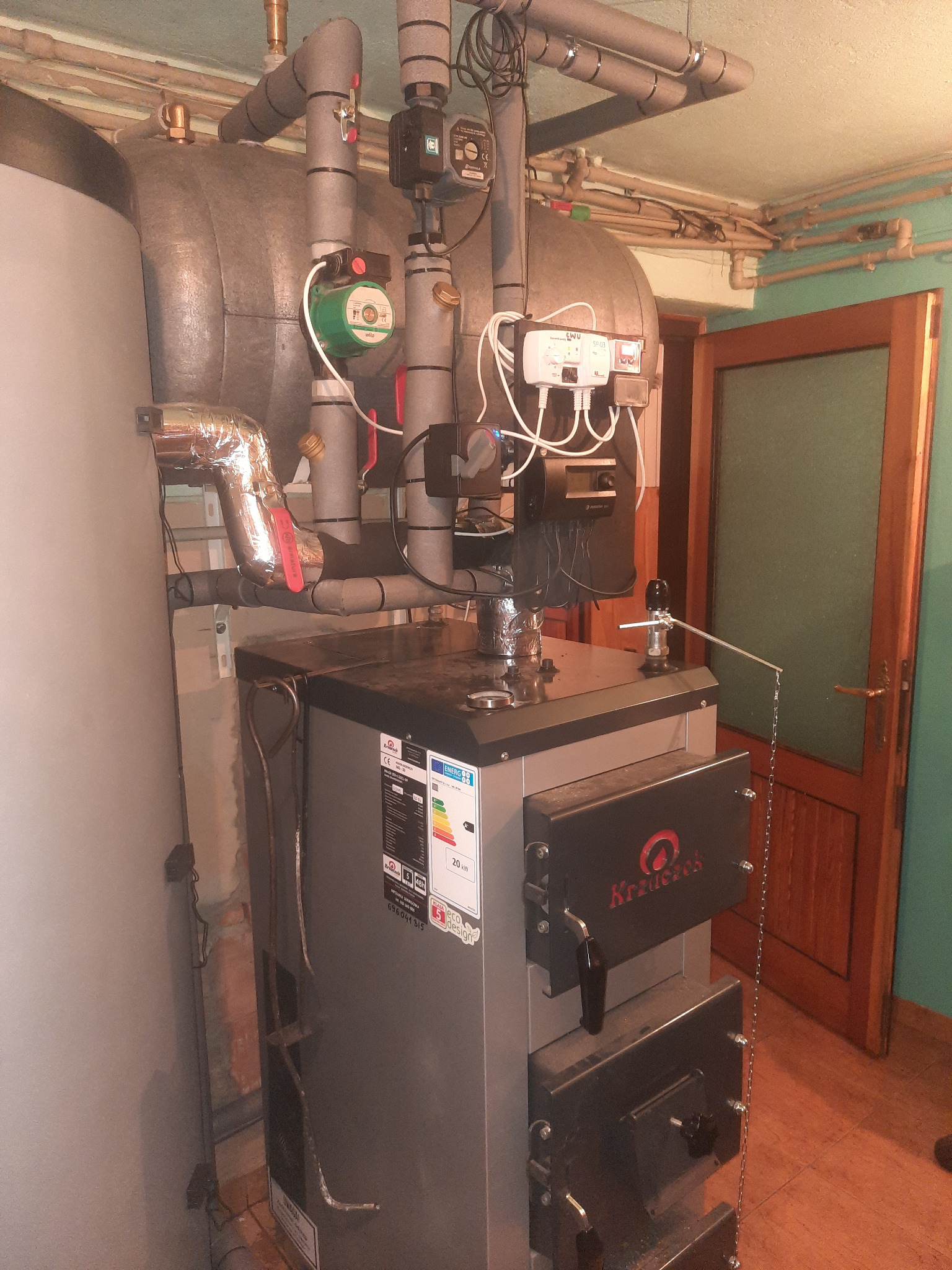Kocioł na paliwo stałe marki Krzaczek w kotłowni, z widocznymi rurami instalacji grzewczej, pompą obiegową Wilo oraz sterownikiem CWU SP-03, z naklejką informacyjną o klasie energetycznej.